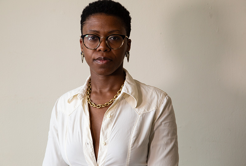 Headshot of Jess Myers, a Black woman with short-cut hair poses in front of a blank beige wall wearing tortiseshell eyeglasses, a gold chain necklace, and a white button blouse.